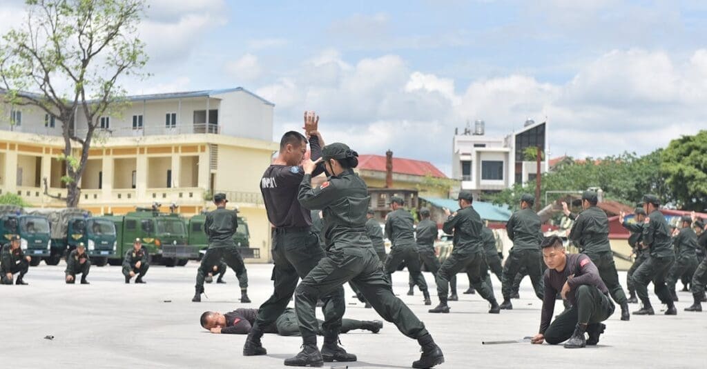 A large group of military or security personnel in uniform conducting outdoor self defense training for beginners, practicing hand-to-hand combat and disarming techniques.