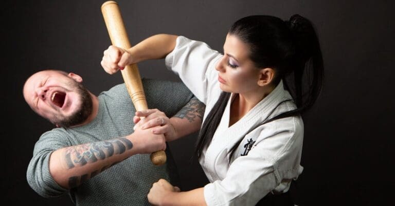 A woman in a martial arts uniform demonstrating a weapon disarm technique against a man with a baseball bat during self defense training for beginners.